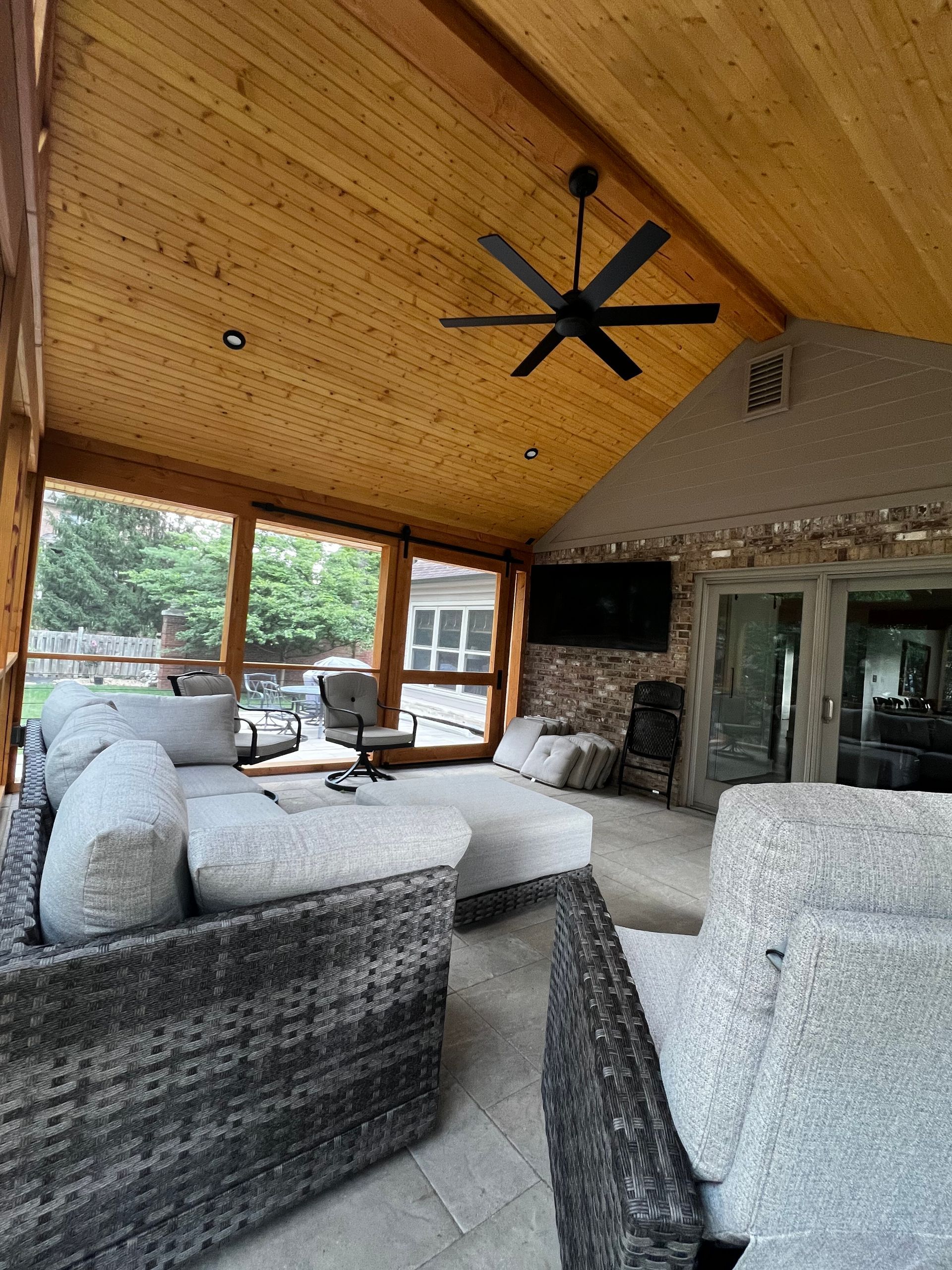 Covered patio with wicker seating, stone fireplace, wood ceiling, large windows, and a ceiling fan