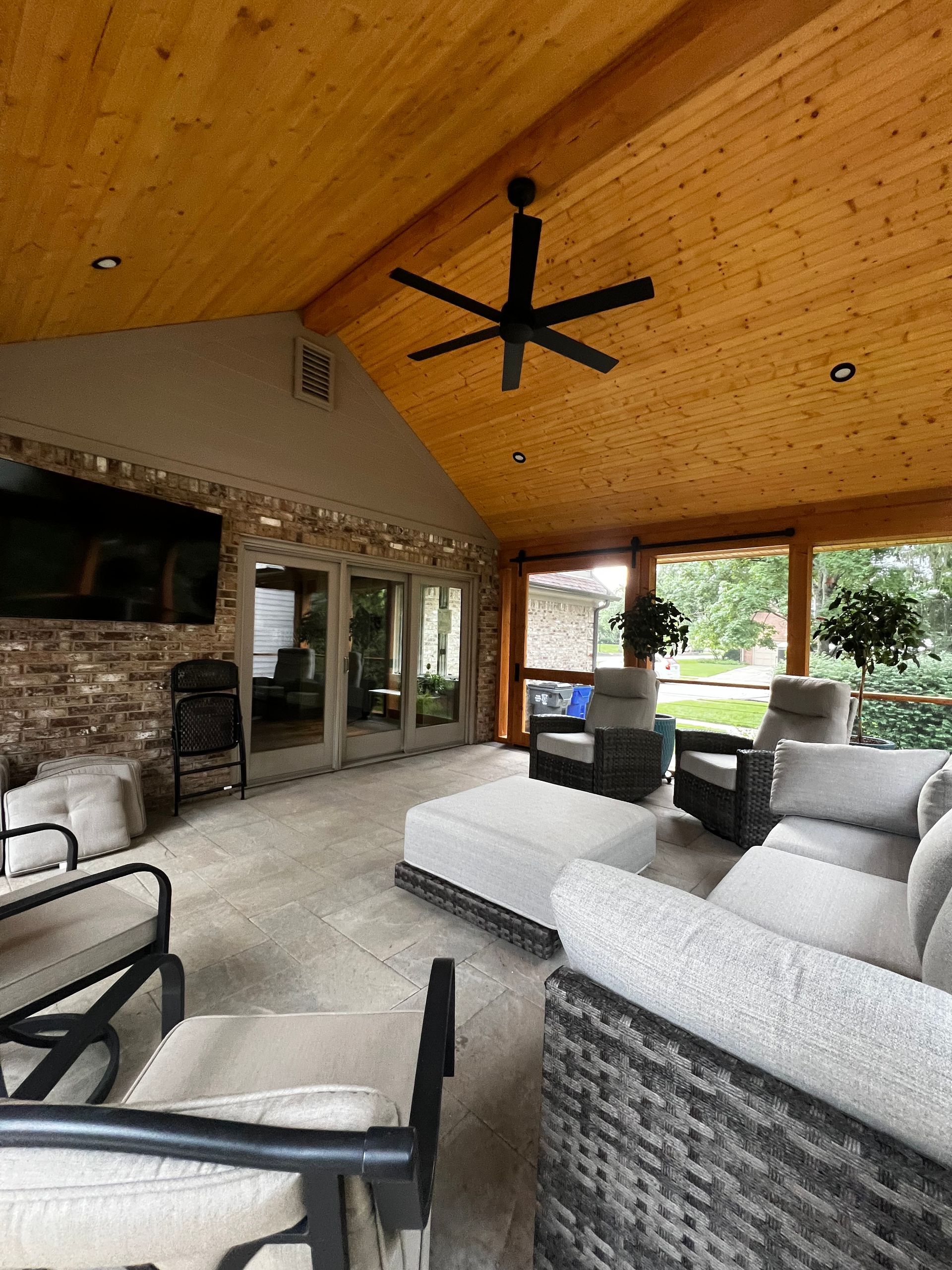 Covered patio with wicker chairs, stone fireplace, and black ceiling fan overlooking a backyard.
