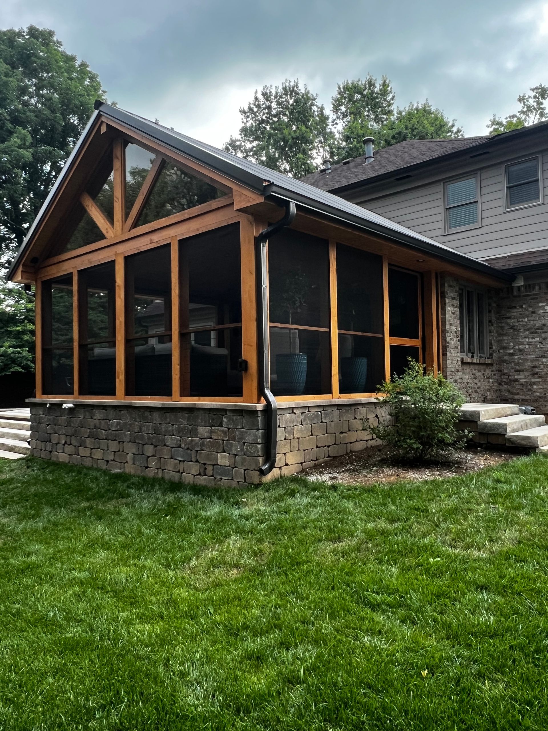 Stone-and-wood screened porch attached to a house, with a grassy yard and trees nearby.