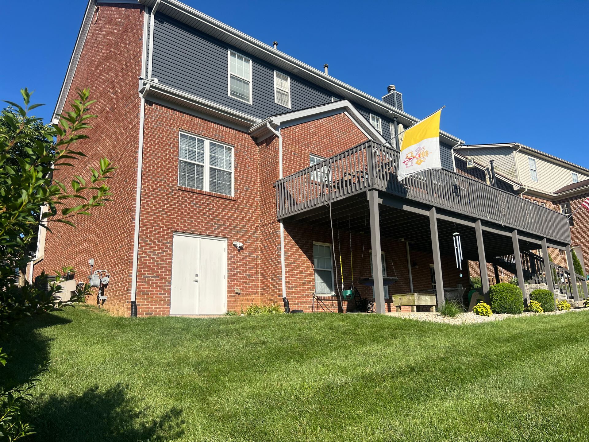 Brick house with dark roof and covered porch on a grassy slope under a clear blue sky