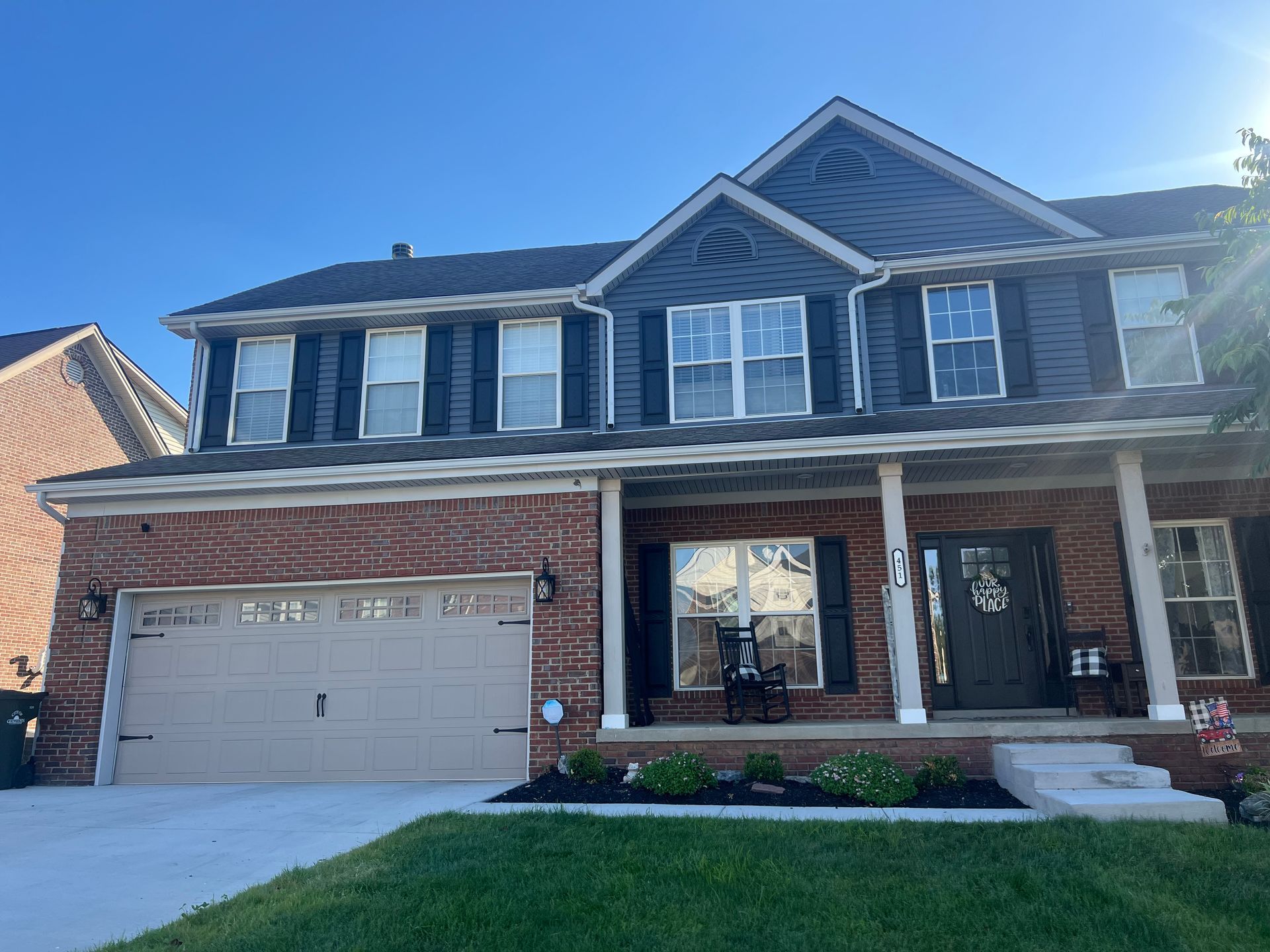 Two-story brick suburban house with a two-car garage, front porch, and landscaped lawn in daylight