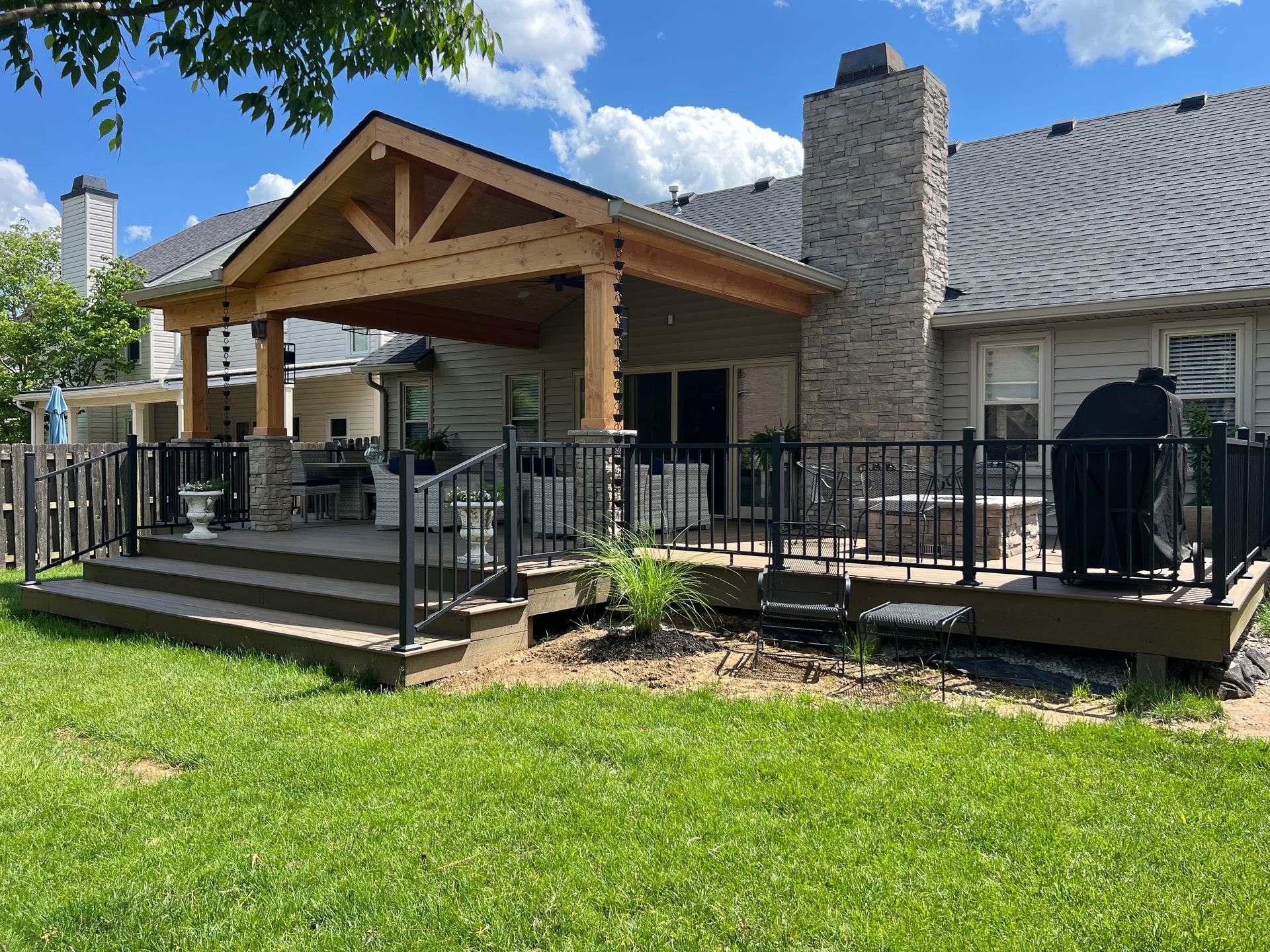 Back deck and covered patio on a stone house, with black railings and green lawn under a blue sky