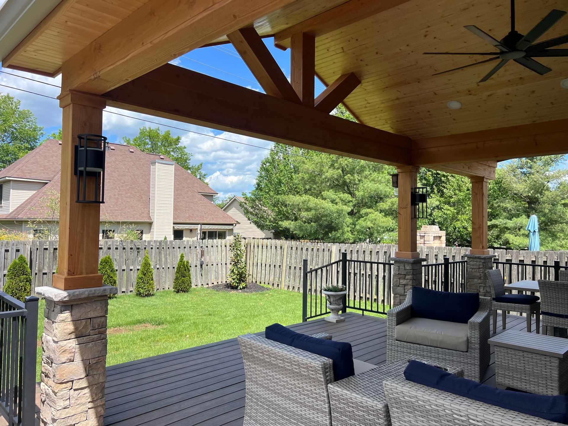 Covered patio with wicker seating, wood ceiling fan, and a green yard with wooden fence behind it