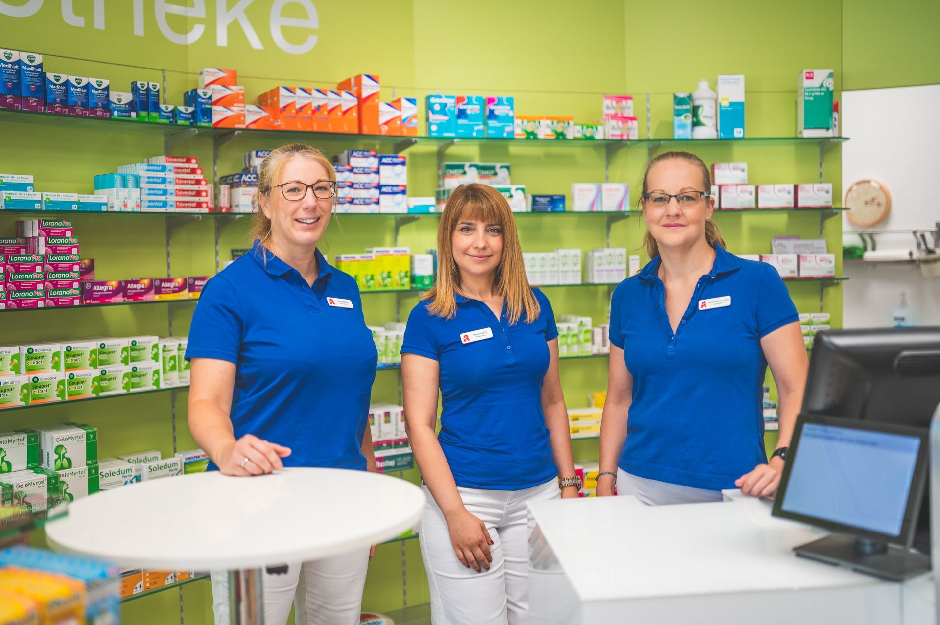 Drei Angestellte in blauen Uniformen stehen an einem Apothekentresen, hinter ihnen befinden sich Regale mit Produkten.