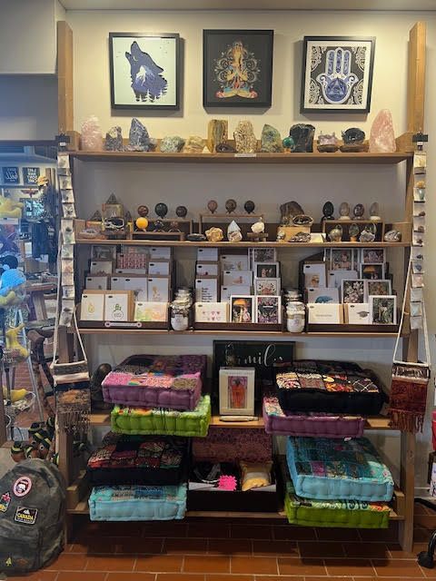 A wooden store display shelf holds crystals, greeting cards, and folded textile cushions, topped by three framed artworks.