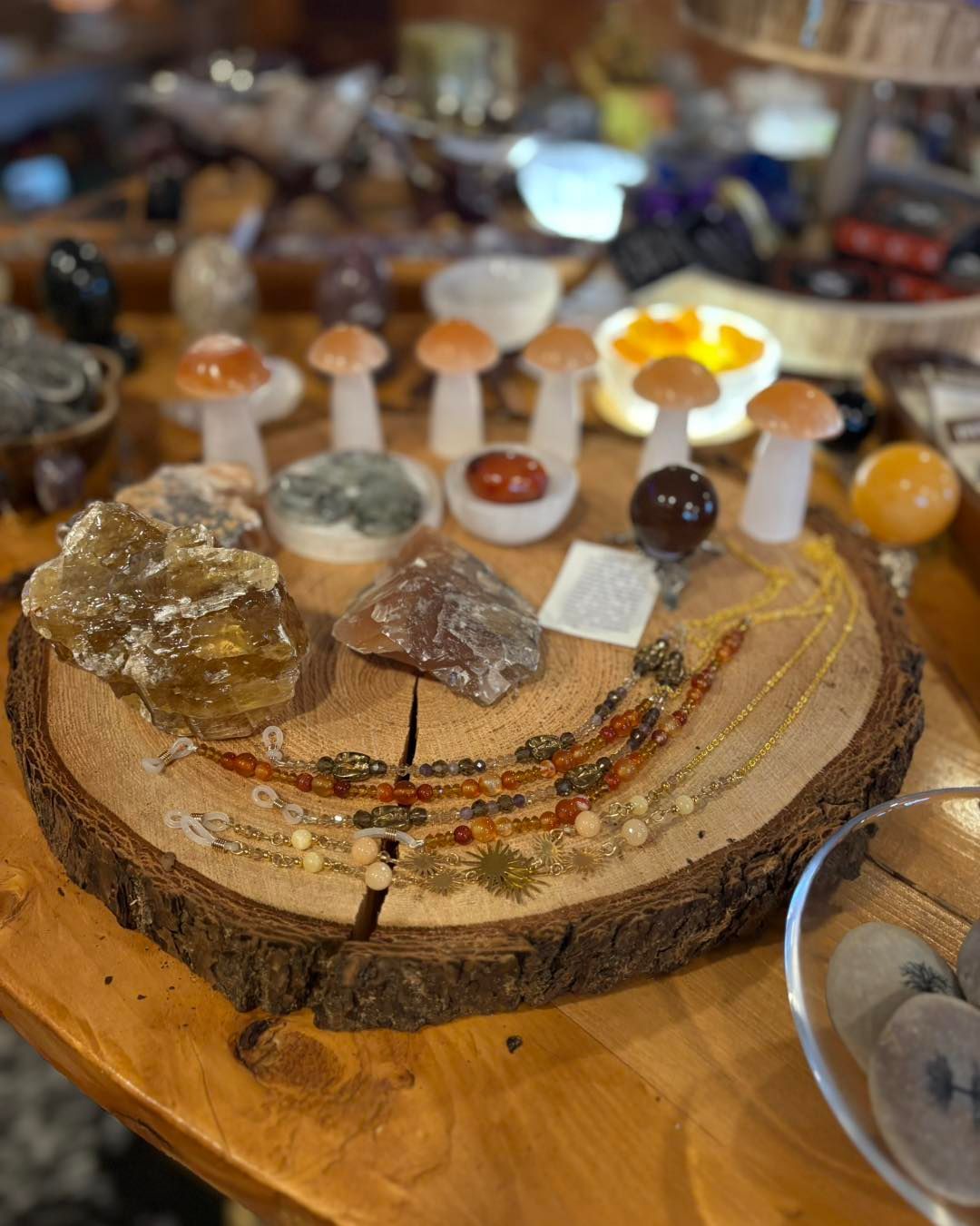 A wood slice display featuring crystal mushrooms, raw stones, a sphere, and beaded necklaces on a wooden table.