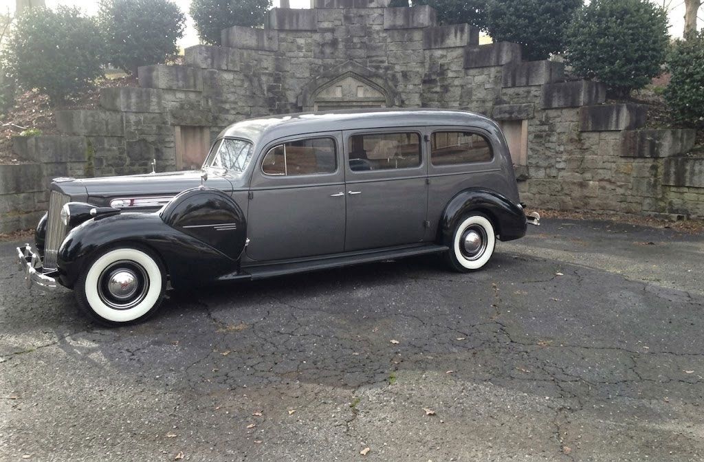 Vintage gray classic car parked on pavement beside a stone wall, viewed from the side.