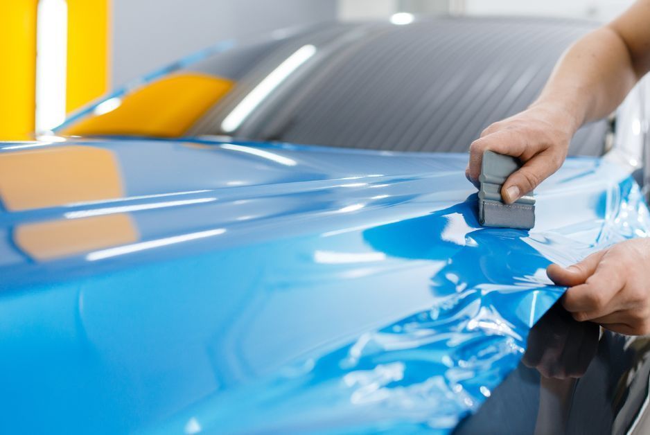 Person applying blue vinyl wrap to a car hood using a squeegee.