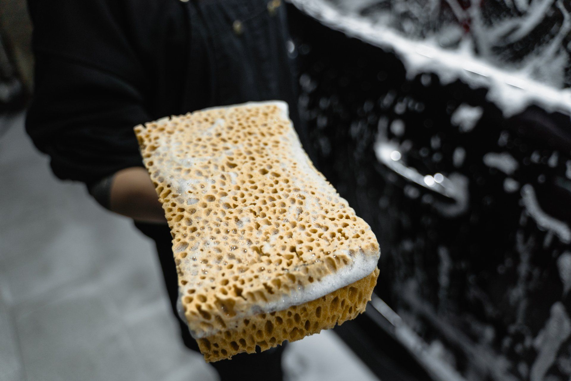 Person holding a wet, soapy sponge next to a black car door, washing the vehicle.