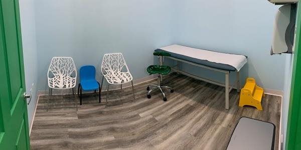 Small exam room with a blue exam table, two white chairs, a stool, and a yellow step stool