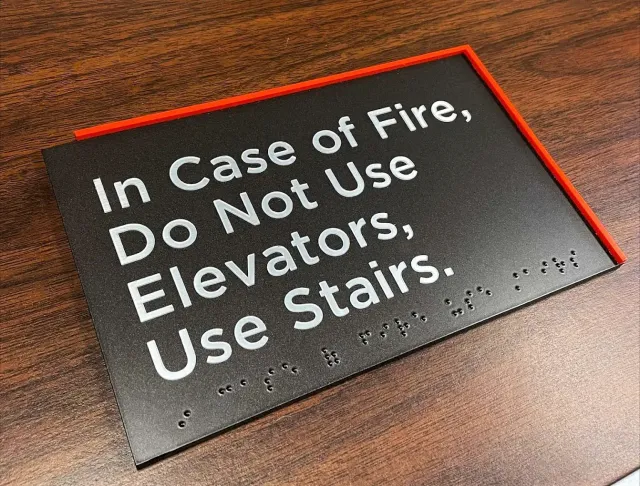 Sign: "In Case of Fire, Do Not Use Elevators, Use Stairs." Includes Braille, red border, on a wooden surface.