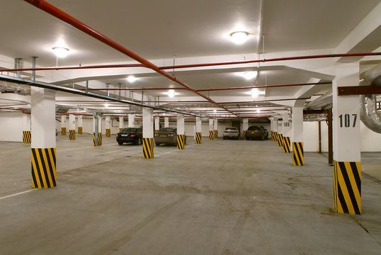 Indoor parking garage with cars. Yellow and black striped columns, concrete floor, and overhead pipes.