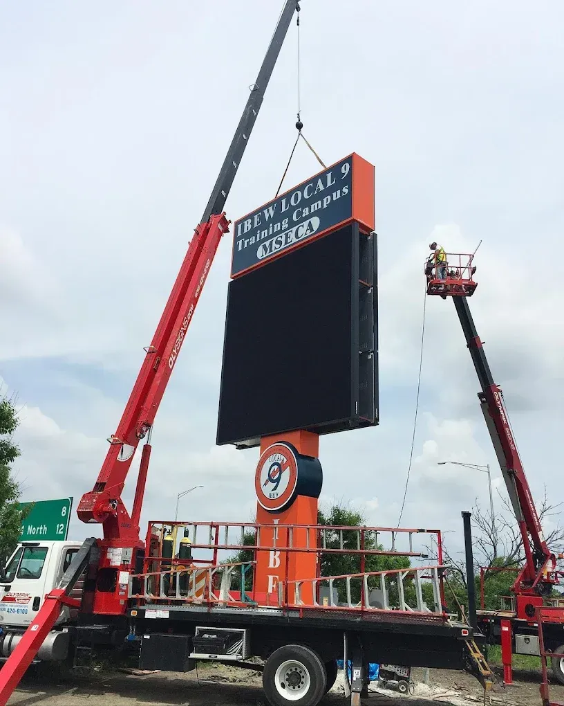 Two cranes lift a new LED sign for IBEW Local 4 Training Campus. Orange and black colors. Two workers on lifts.