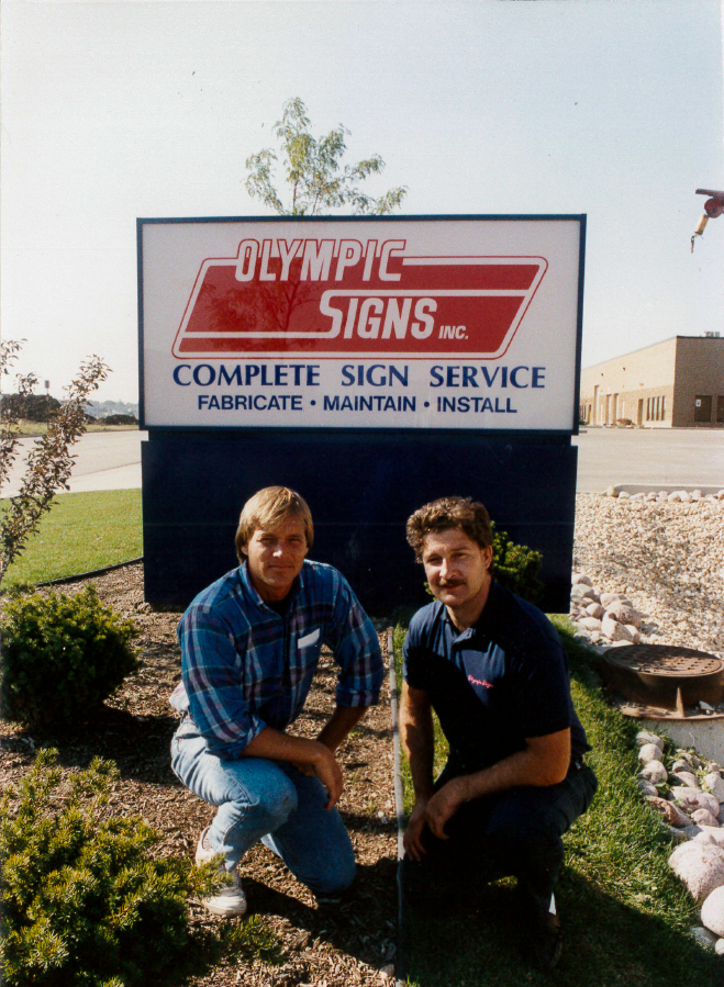 Two men kneel in front of a sign.