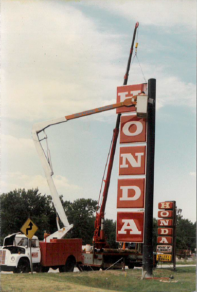 A cherry picker lifts a Honda sign panel. Red and white letters against a brown pole. Parked truck and roadside trees.