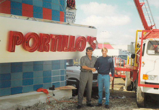 Two men shake hands in front of a "Portillo's" sign, with a truck crane nearby.