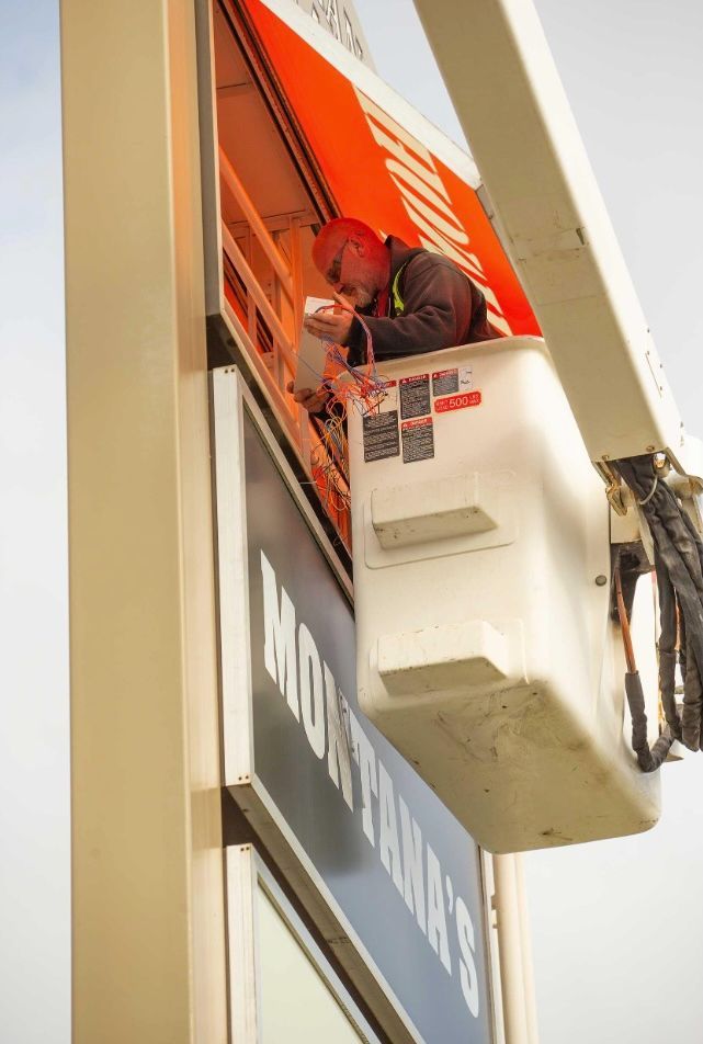 Sign being installed by crane and lift truck. IBEW Local 4 training campus. Worker in lift. Orange and blue.