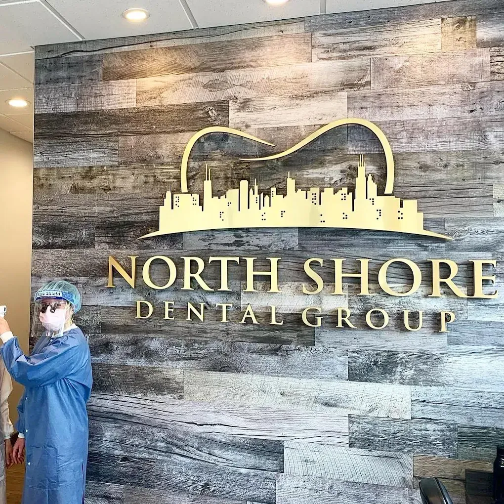 Dentist in protective gear standing near North Shore Dental Group logo on a wooden wall.