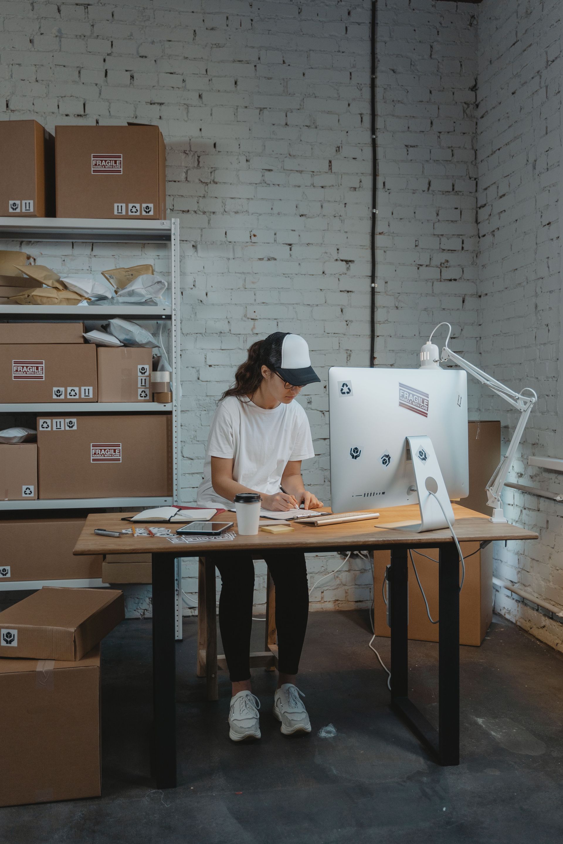 Person working at a desk in a brick-walled office, with shelves of boxes and a large monitor.