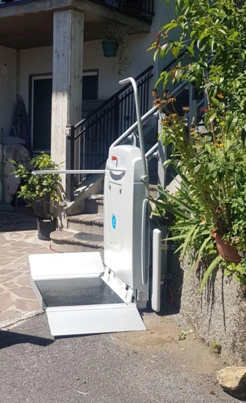 Montascale esterno per sedie a rotelle con rampa, situato accanto all'ingresso di un edificio e circondato da piante in vaso.
