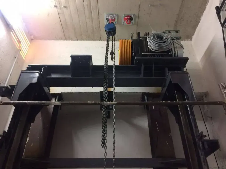 Paranco industriale con catena e motore montato su una trave del soffitto in un interno simile a un magazzino.