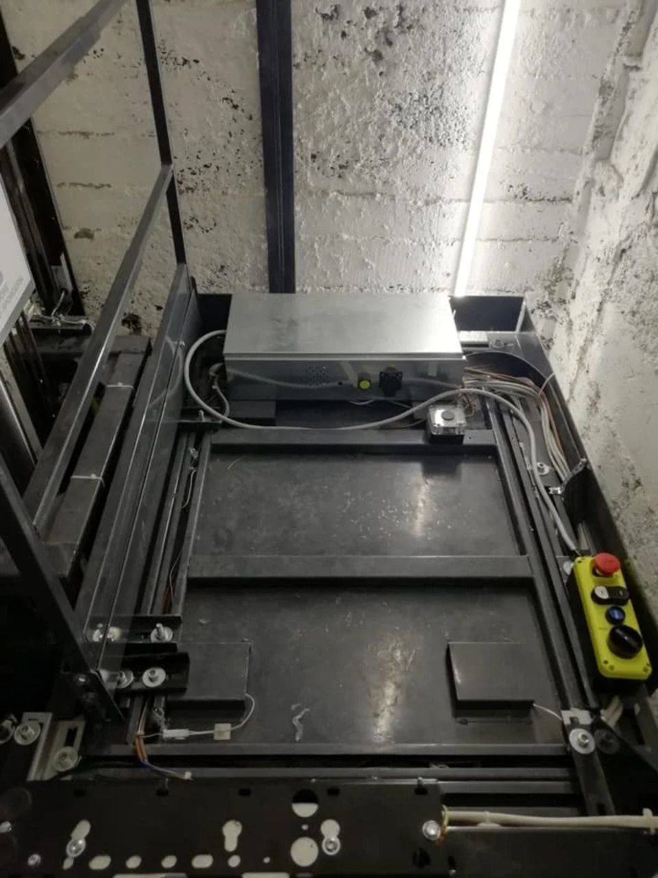 Interno del vano ascensore industriale con macchinari neri, cavi e un pannello di controllo giallo sulla destra.