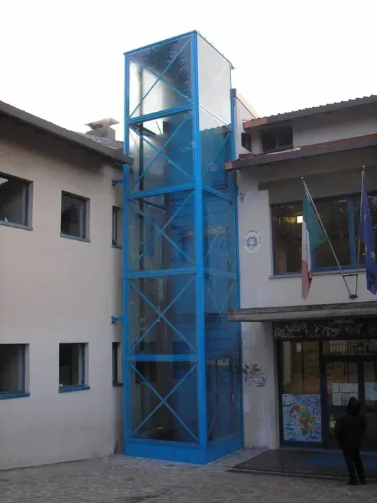 Torre ascensore in vetro blu annessa al cortile di un edificio, con una persona in piedi nelle vicinanze.