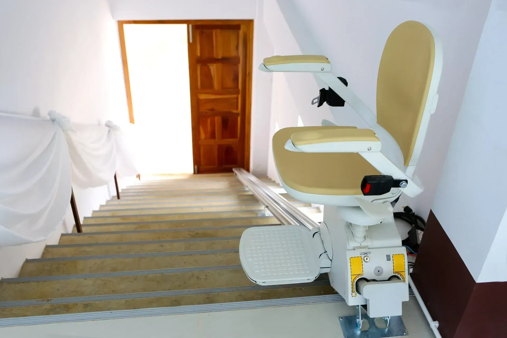 Montascale su una scala interna con sedile bianco e corrimano marrone, vicino a una porta di legno aperta.