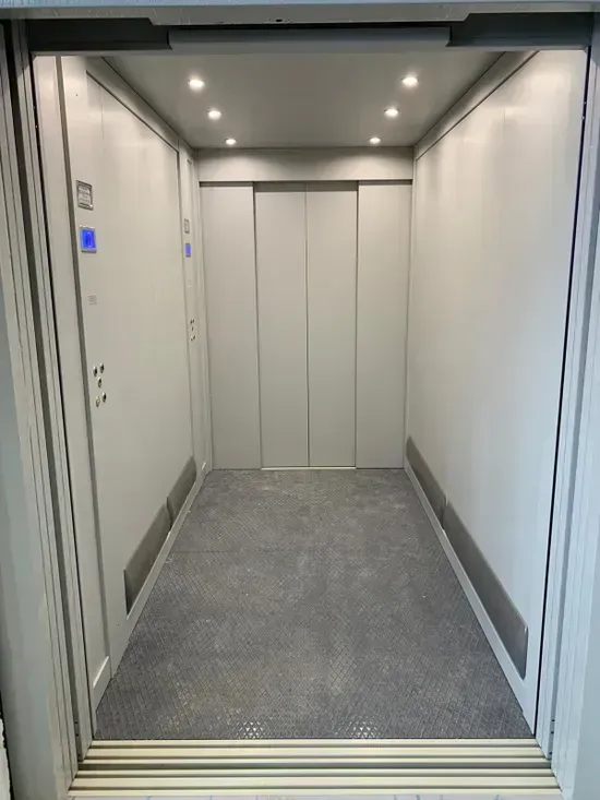 Interno vuoto di un ascensore moderno con pareti argentate, pavimento grigio e porte chiuse.