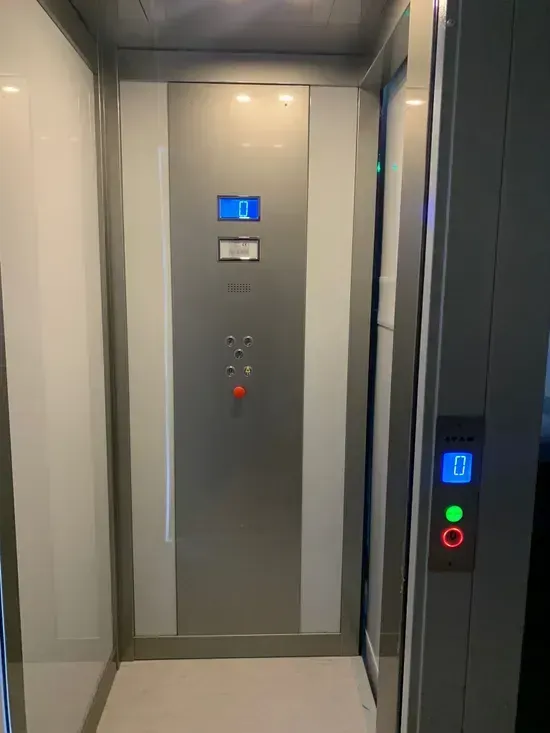 Interno dell'ascensore con pannello di controllo, display blu e pulsante di emergenza rosso a parete.