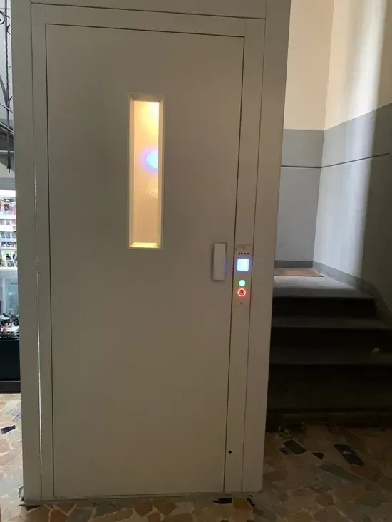 Porta dell'ascensore grigia con finestra stretta e pannello di controllo illuminato nel corridoio di un edificio.
