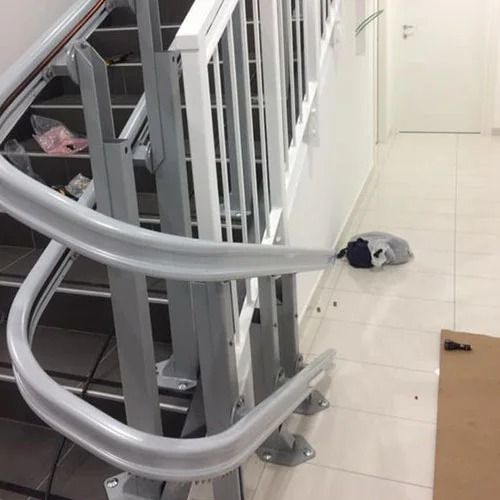 Montascale grigio lungo una scala interna con ringhiere bianche, con una piccola borsa sul pavimento piastrellato.