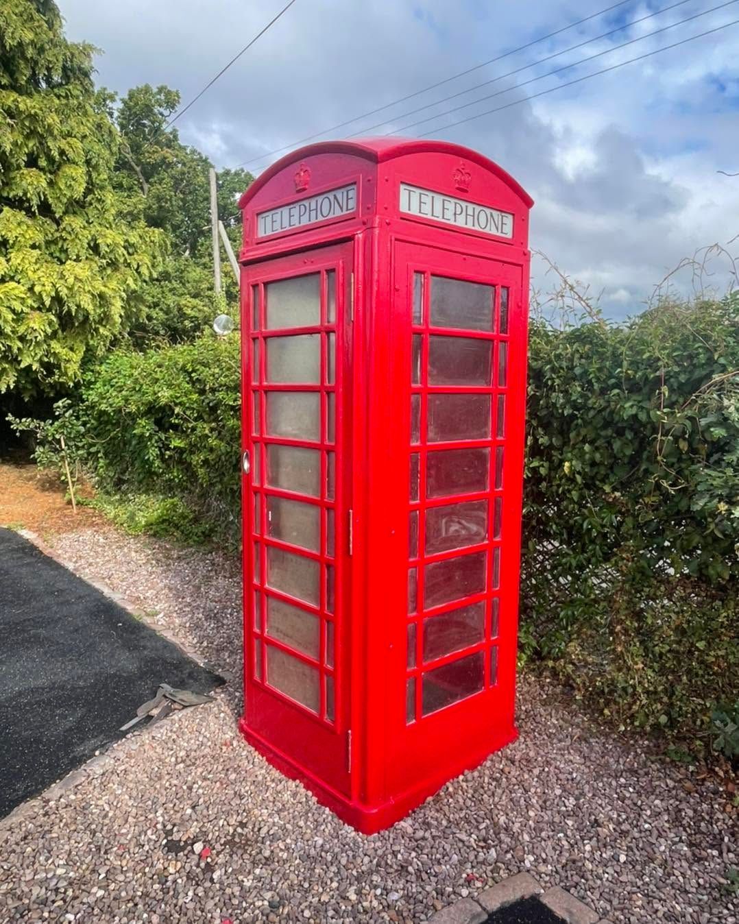 Bright red British-style telephone booth on gravel beside greenery under a cloudy sky