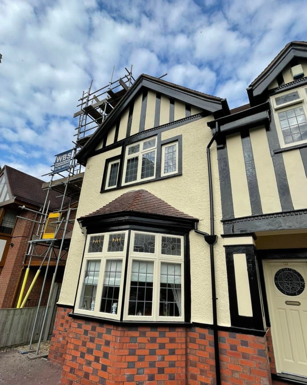 Tudor-style house with cream and black timbering, red brick base, and a bay window under a cloudy sky.