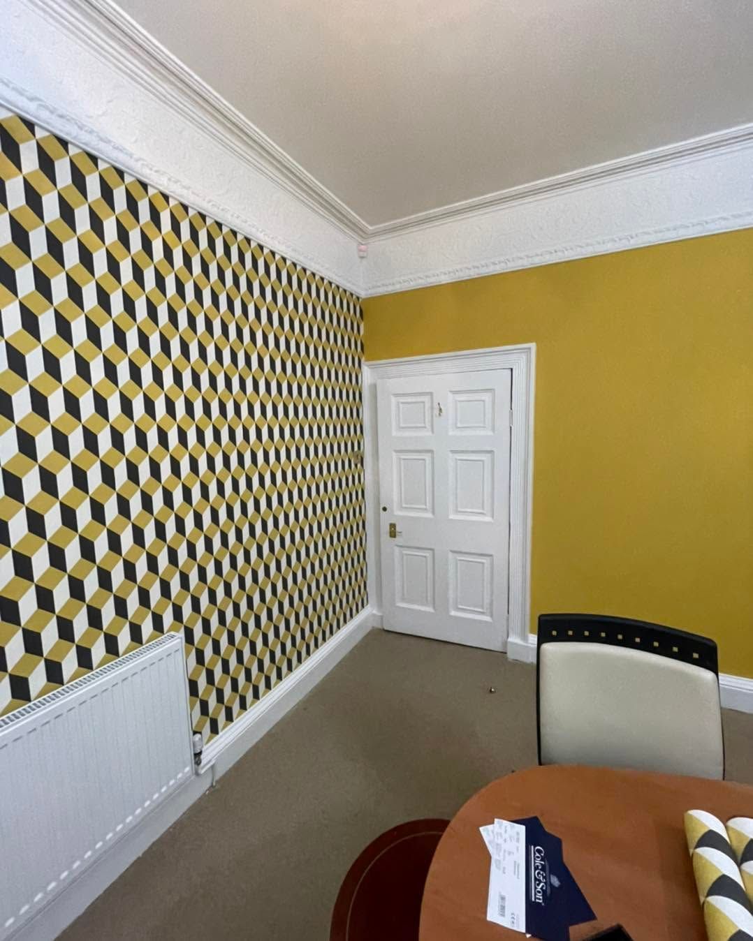 Small room with yellow walls, geometric wallpaper, white door, radiator, and a table in the foreground.