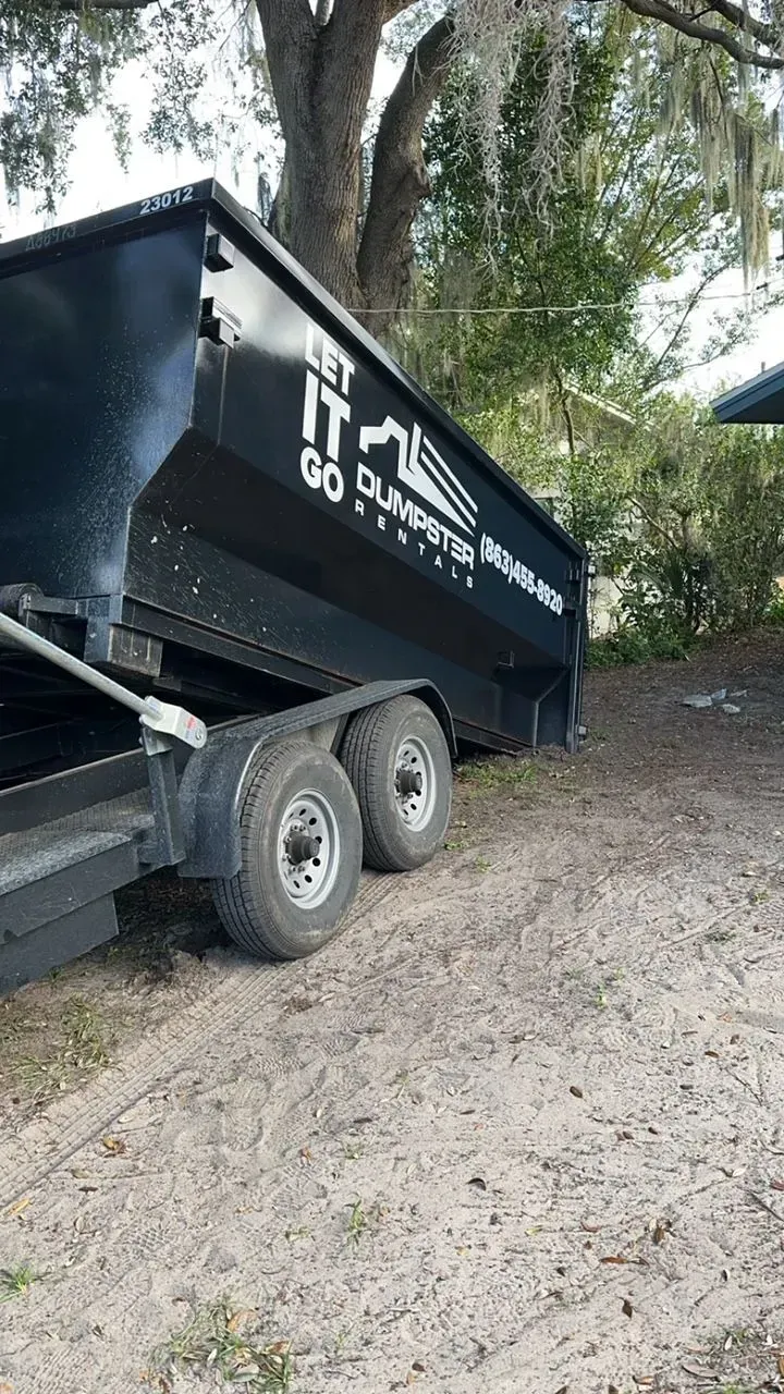Black dump trailer with white lettering parked on a gravel path under trees