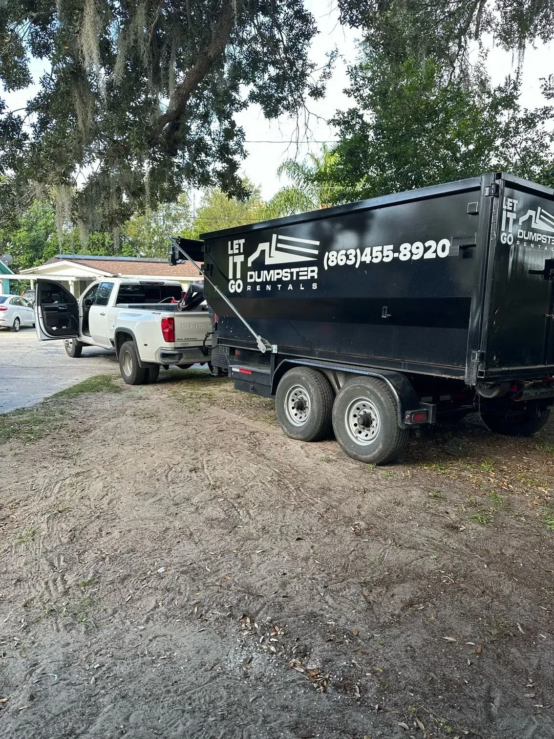 White pickup truck towing a black utility trailer parked under trees on a dirt drive
