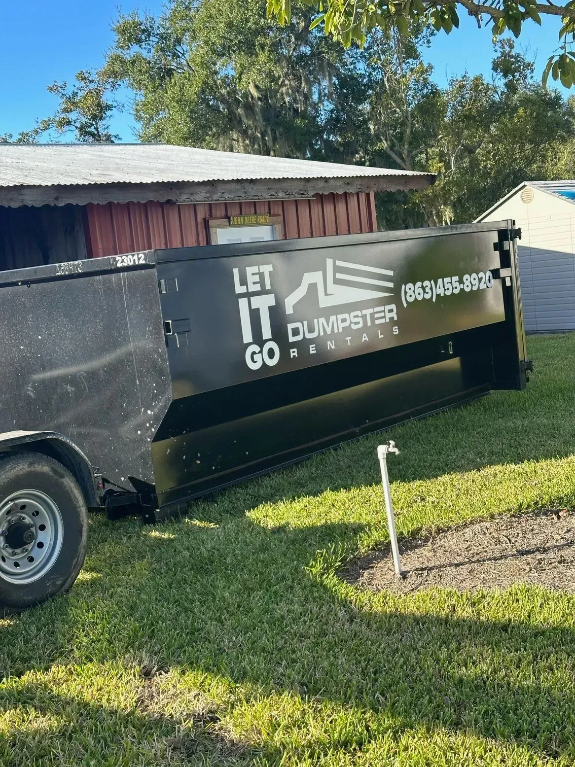 Black dumpster trailer with “LET IT GO” branding parked on grass beside a house in sunlight