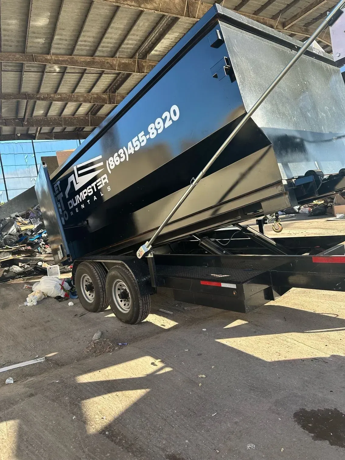 Black dump trailer tipped on an angle under a metal awning, with debris and trash nearby.