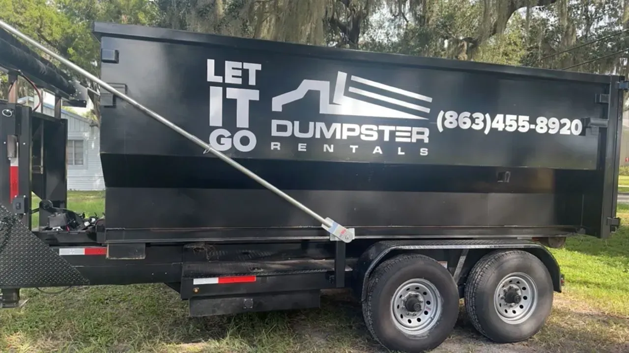 Black dumpster rental trailer with “LET IT GO” logo, parked on grass near trees.