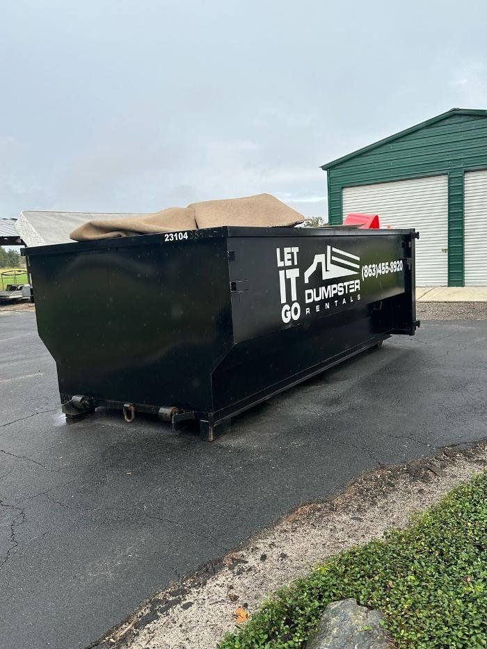 Black construction dumpster with open top beside a green building on a wet lot