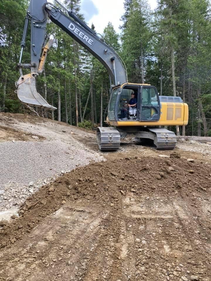 Excavatrice jaune John Deere creusant la terre dans une zone boisée.