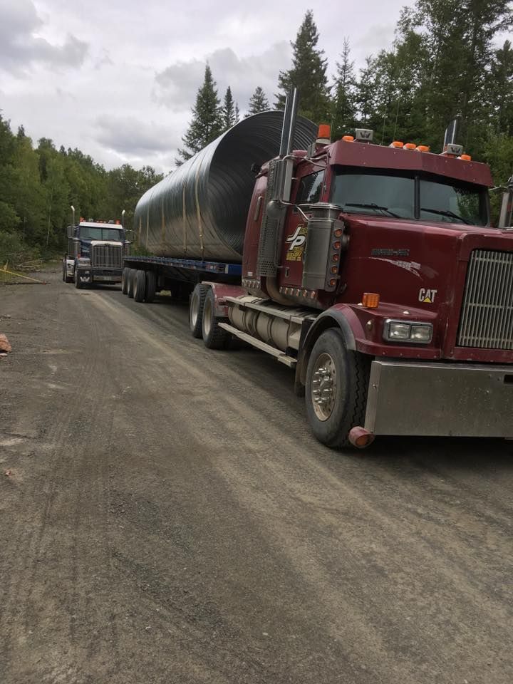 Deux semi-remorques transportant un gros tuyau sur une route de gravier, sur fond d'arbres et de ciel nuageux.