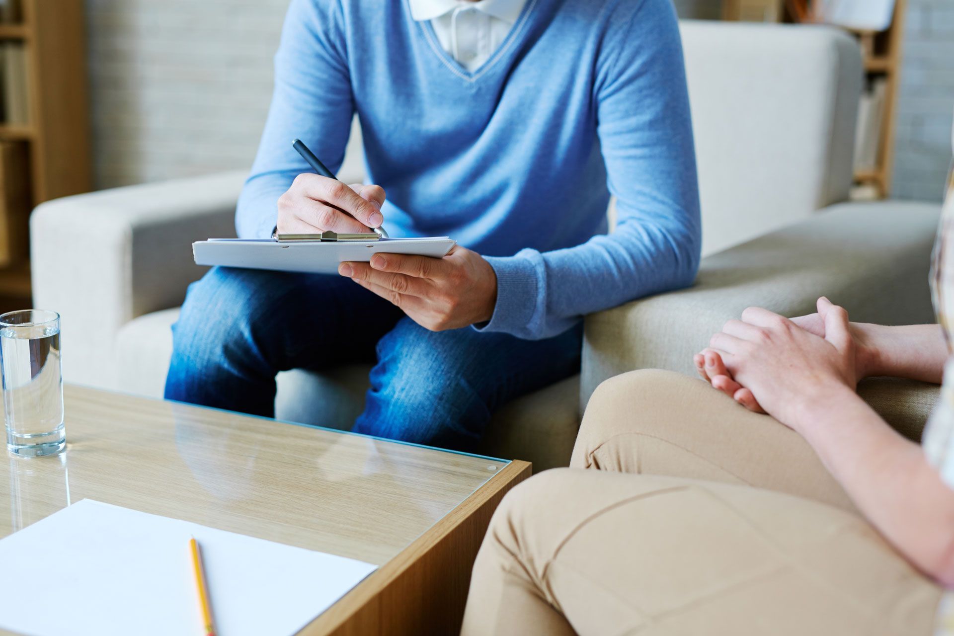 Therapist in blue sweater takes notes while speaking with patient. Beige couch, table, and glass of water.