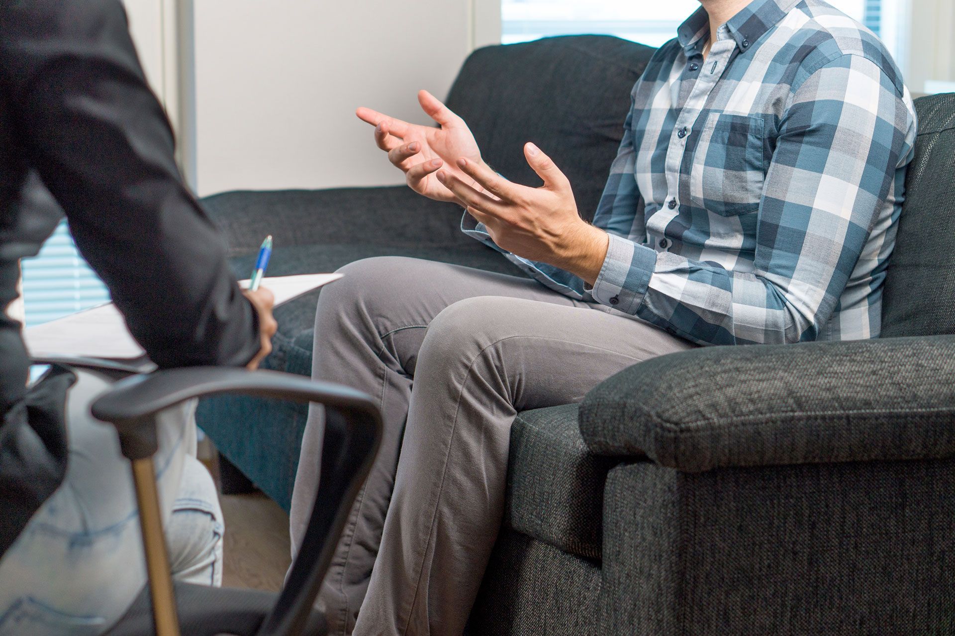 Man in plaid shirt gesturing, talking to person in suit, seated indoors.