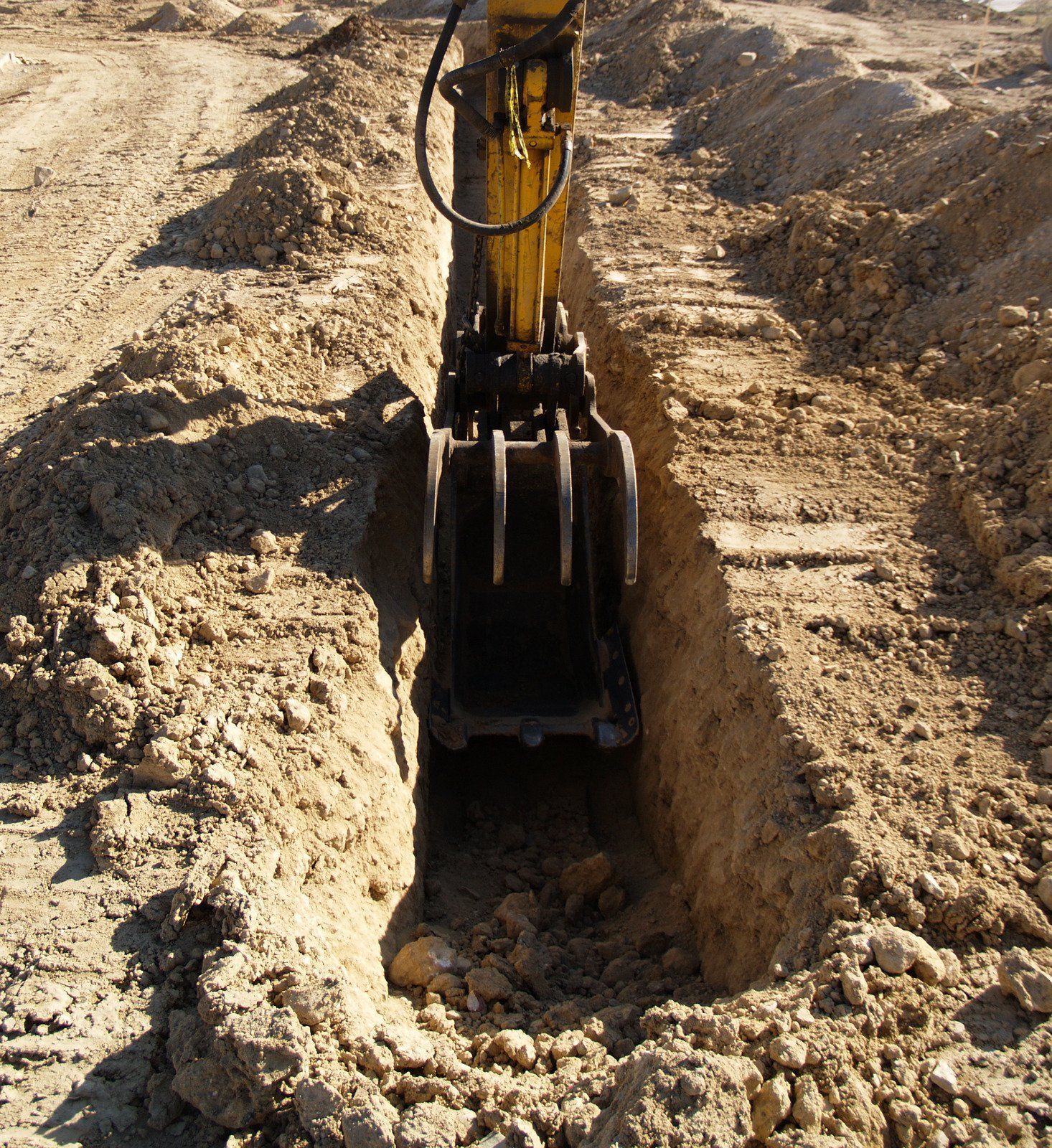 A yellow excavator bucket with prongs digs a deep, narrow trench into the brown dirt of a construction site.