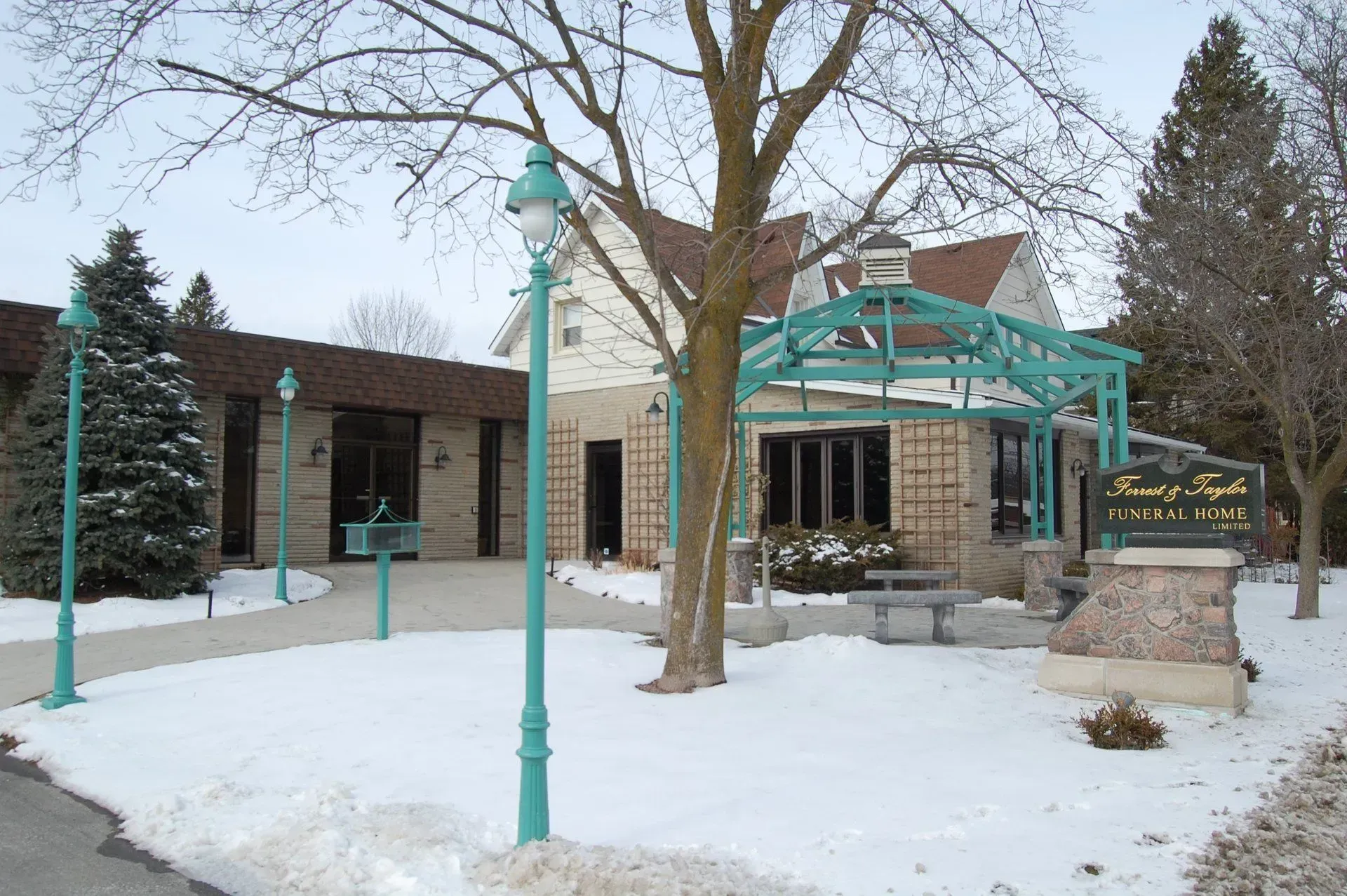 Snowy entrance to a stone building with teal awning and lamp posts, surrounded by leafless trees.