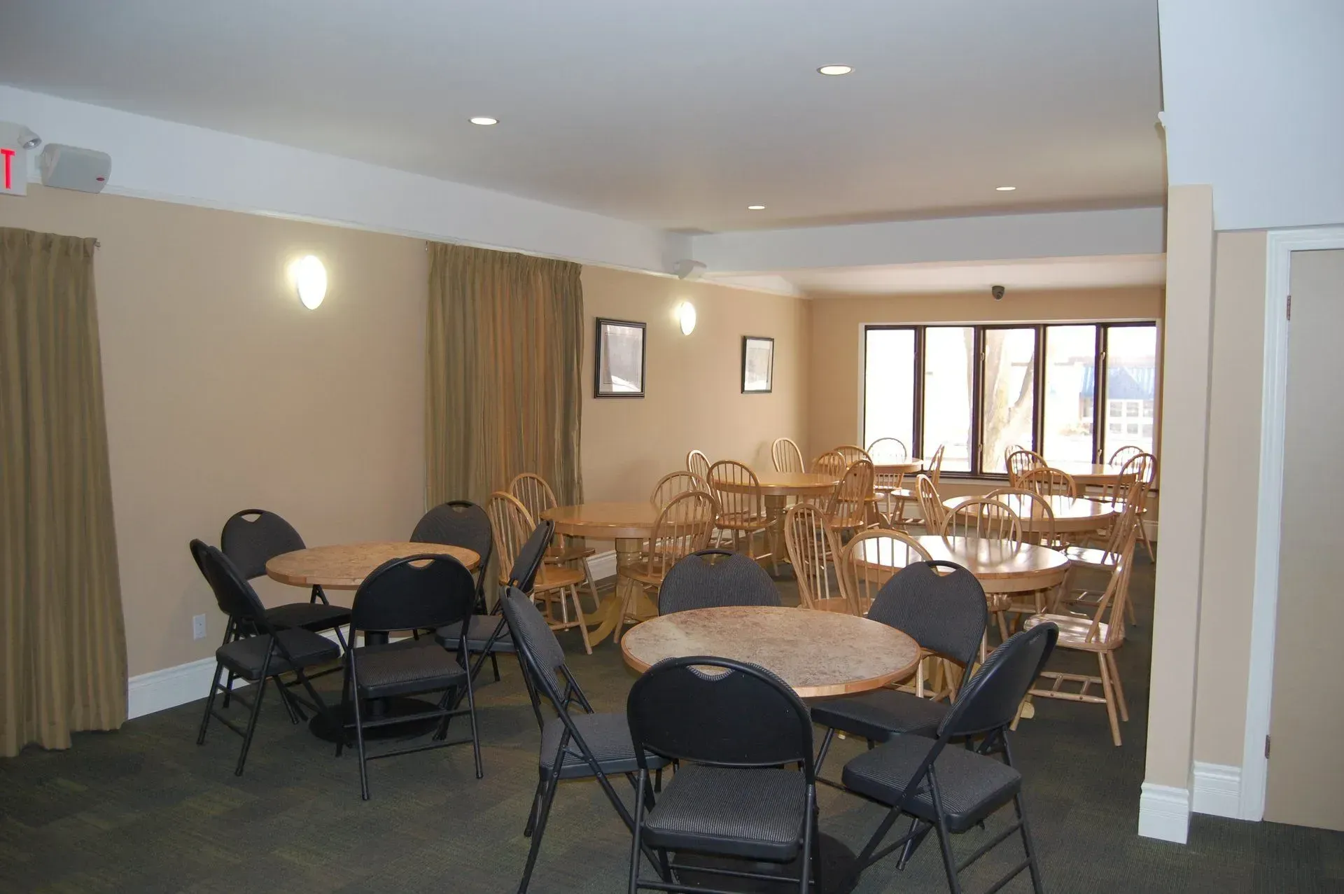 Empty banquet room with round tables, black chairs, beige walls, and windows at the far end