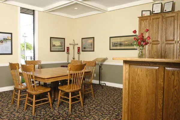 Hotel lobby dining area with wooden tables and chairs, framed art on beige walls, and a reception counter.