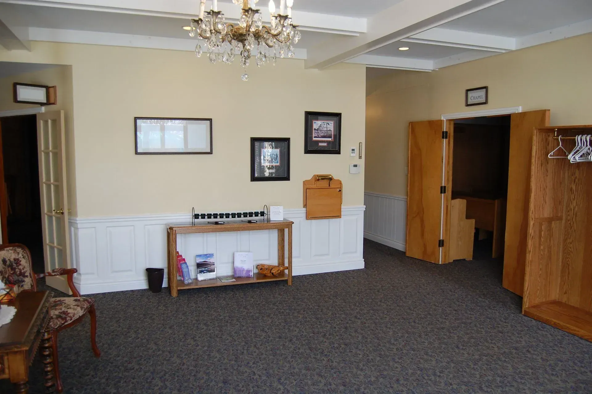 Empty hotel lobby with beige walls, dark carpet, chandelier, framed art, and wooden storage lockers.